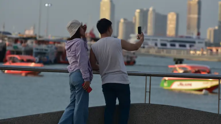 Two tourists taking a selfie by the waterfront with modern skyscrapers and boats in the background, themed around Dubai vs Abu Dhabi.